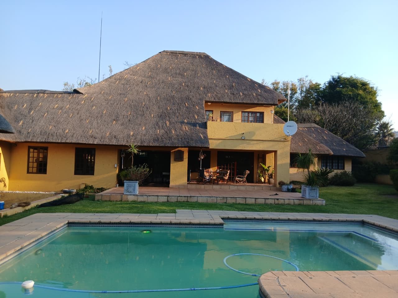 A home with warm yellow walls and a full thatch roof beside a swimming pool under a clear Gauteng sky.