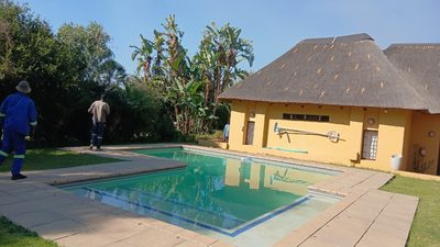 A home with a full thatch roof and a swimming pool, with workers on site by the water.