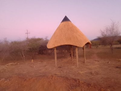 A freshly thatched round rondavel with a pointed conical roof, standing on cleared ground at dusk.